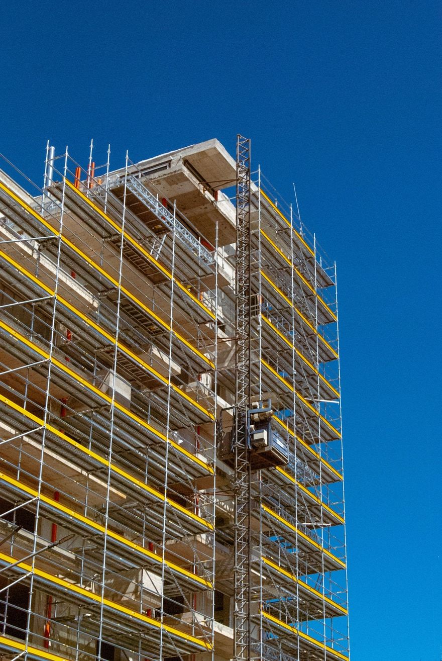 Rohbau eines Gebäudes, rundum eingerüstet mit Systemgerüst und gelben Bordbrettern, vor blauem Himmel.