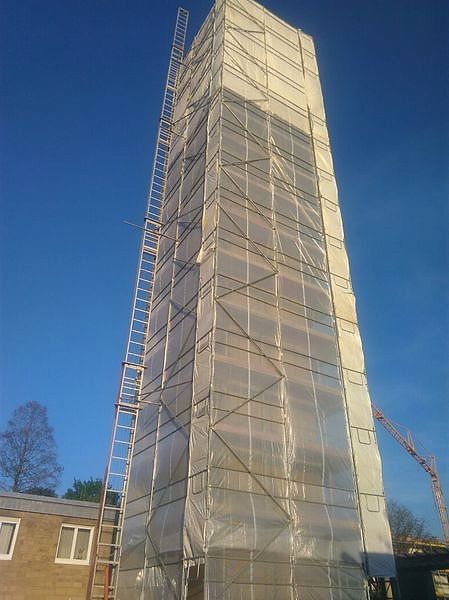 Eingehauster Gerüstturm mit außen geführtem Leiteraufstieg, dahinter ein Baukran.